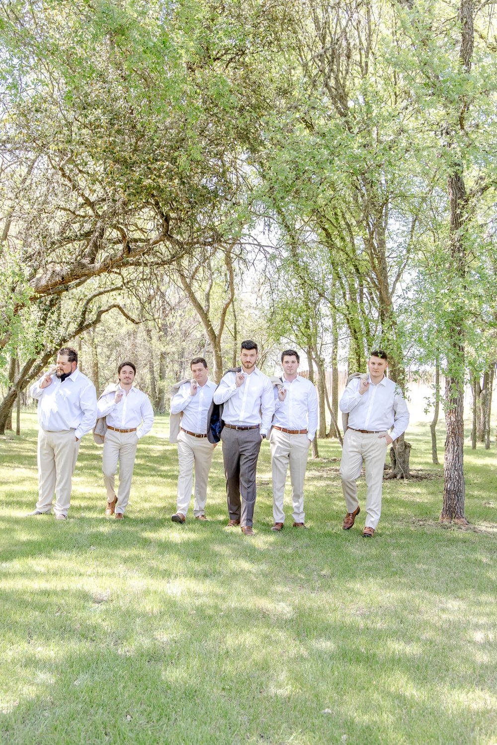 A group of groomsmen in light colored shirts and pants pose casually while standing on grass beneath trees in a park.