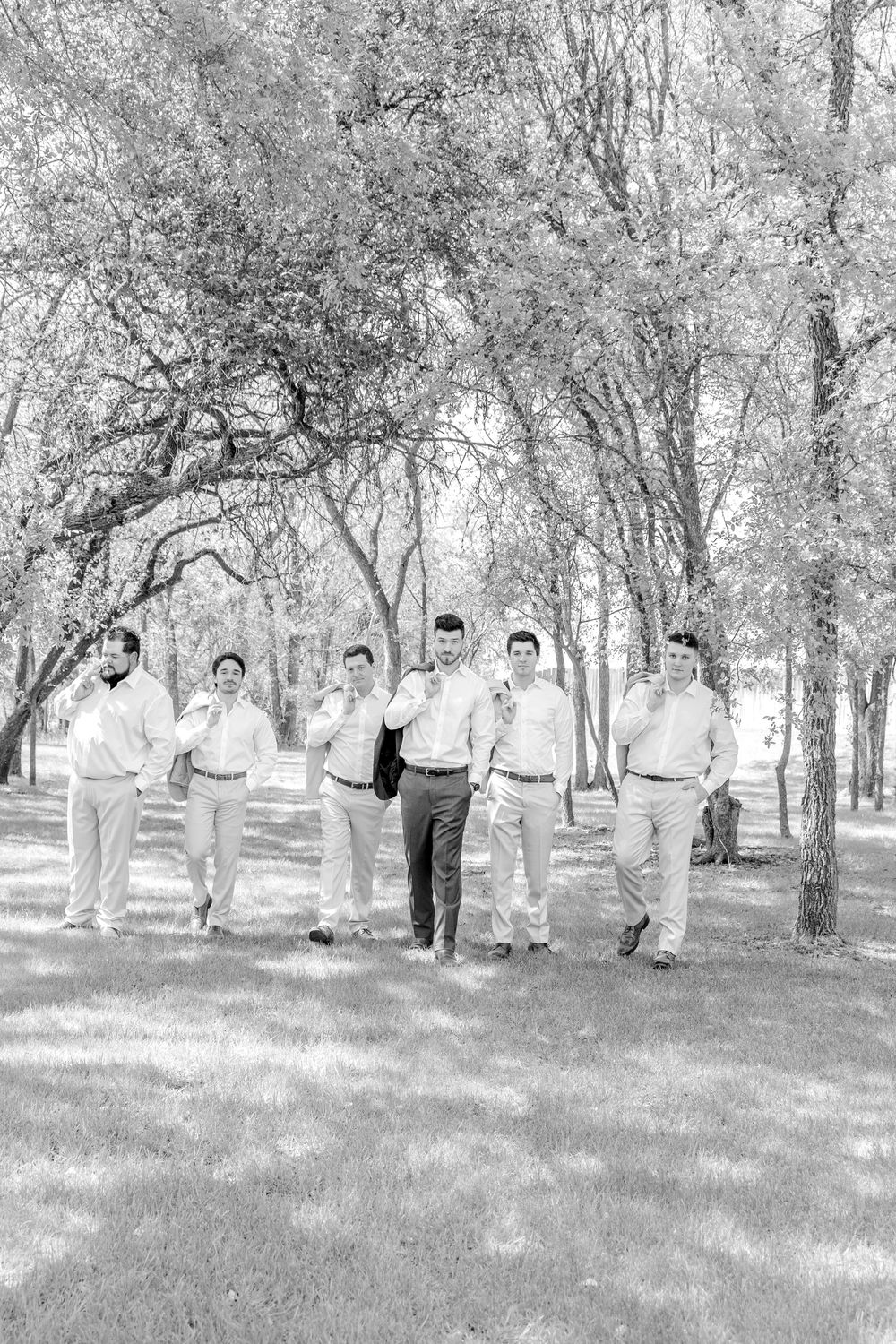 A black and white photograph shows a group of people in light colored attire walking together through a wooded area outdoors.