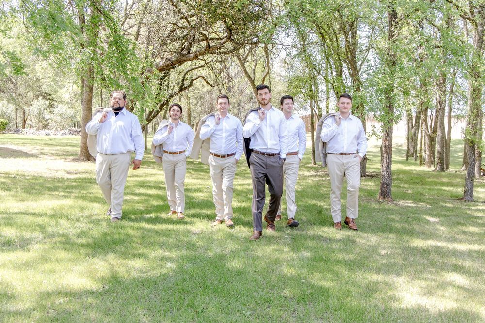 A wedding party in light grey and white attire walking together through a sunlit wooded park area.