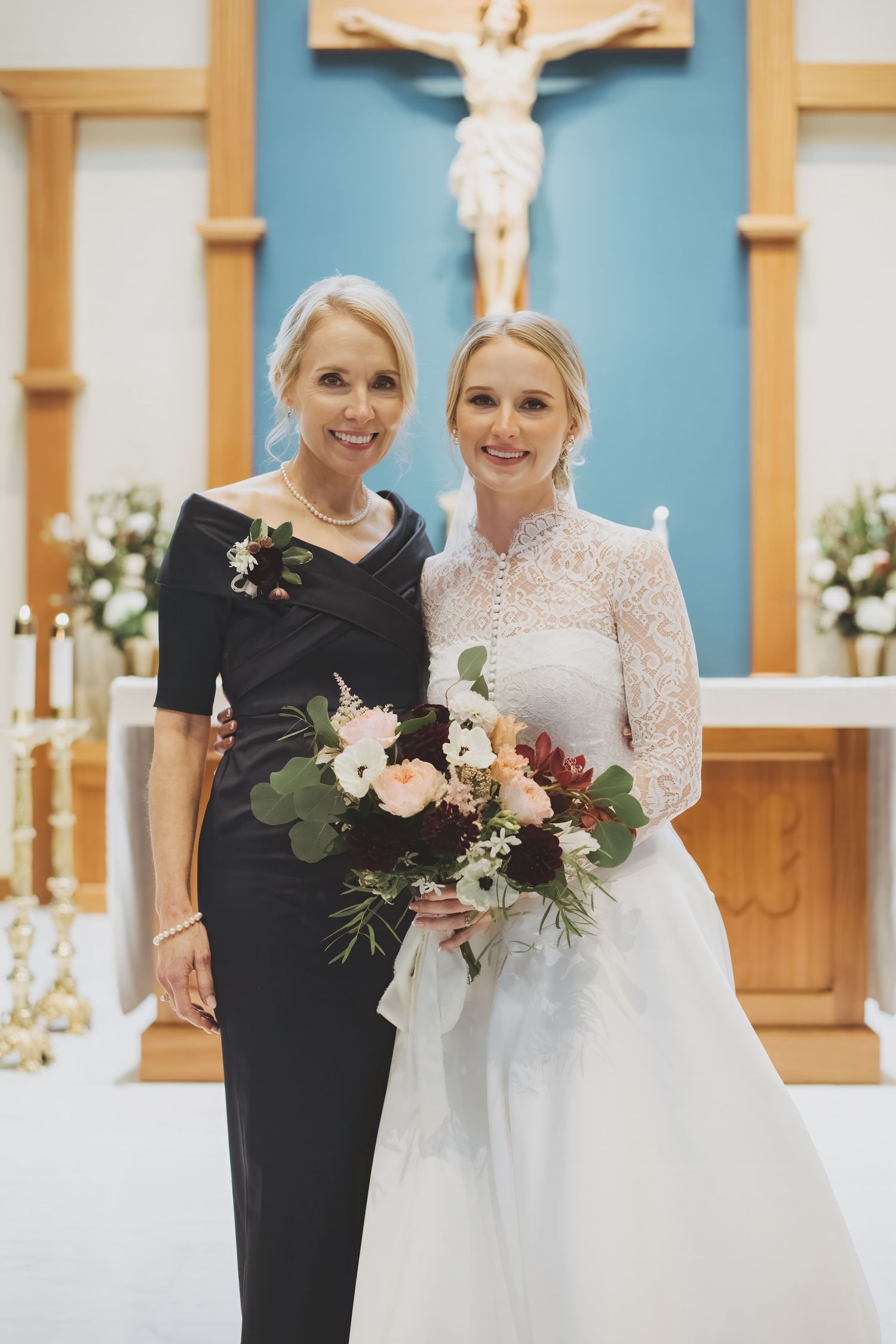 Bride and mother of the bride pose with elegant bridal bouquet featuring peach and white flowers against blue wall backdrop.