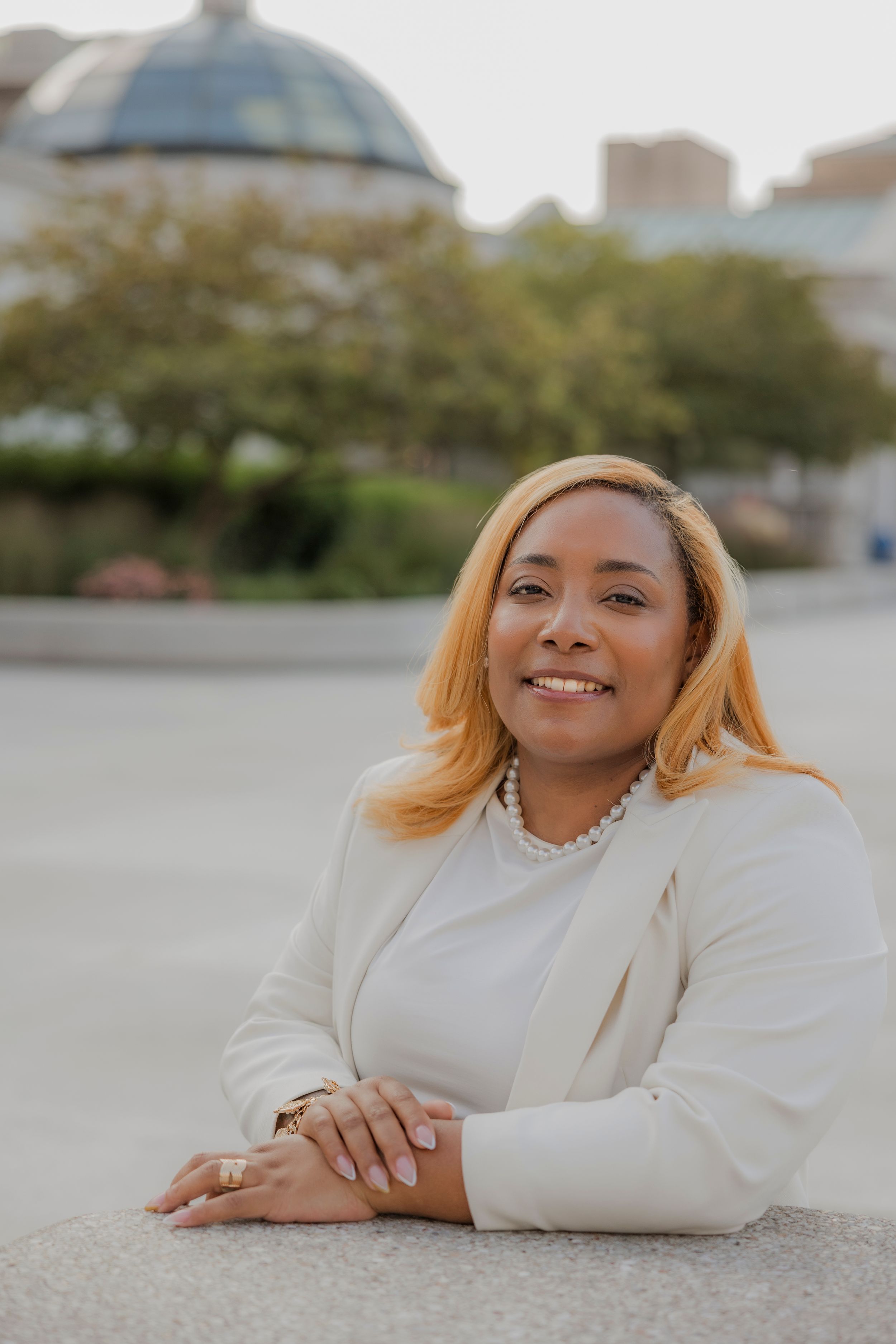 Professional headshot of a person in a white blazer smiling warmly outdoors.