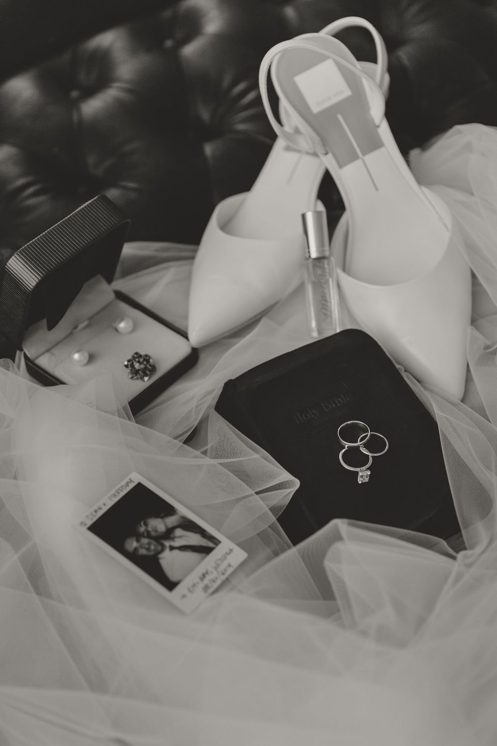Wedding details flatlay with white heels, rings, and photographs on tulle fabric.