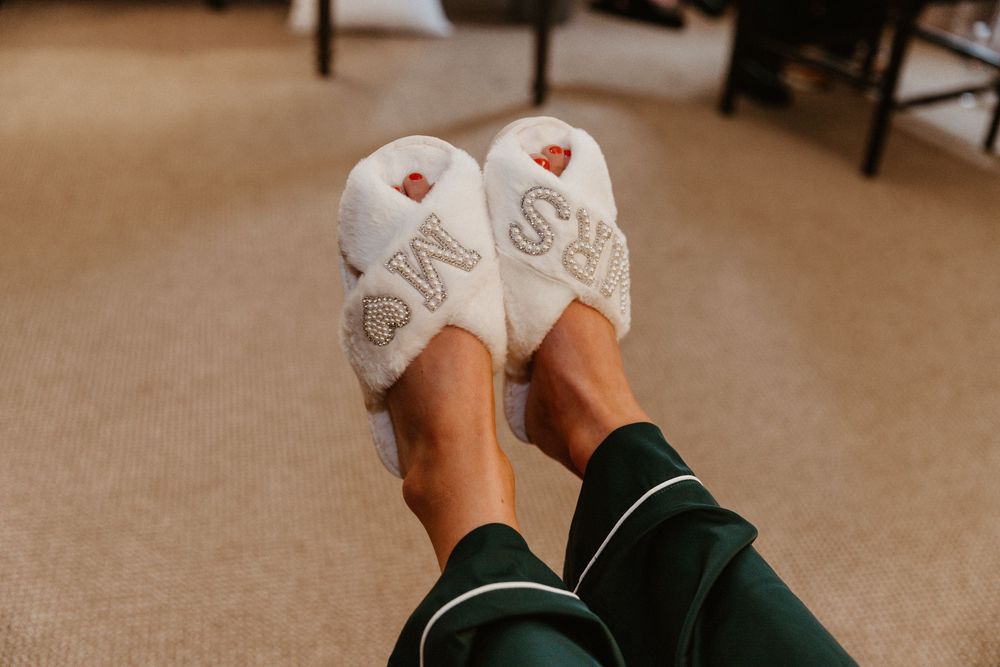 White bridal slippers with 'Mrs' text photographed on beige carpet.