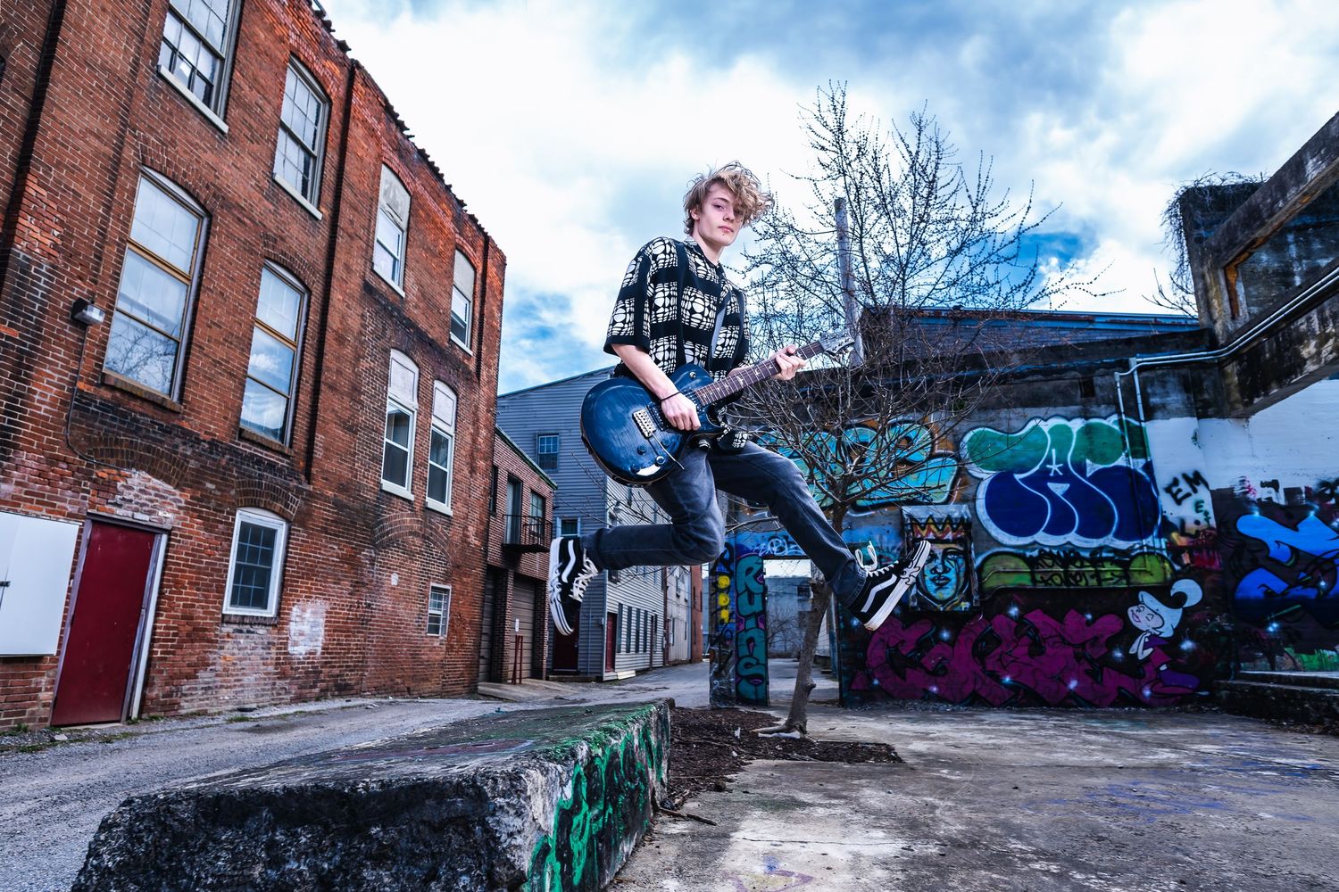 Urban street scene with guitarist jumping in front of graffiti wall and brick buildings under blue sky.