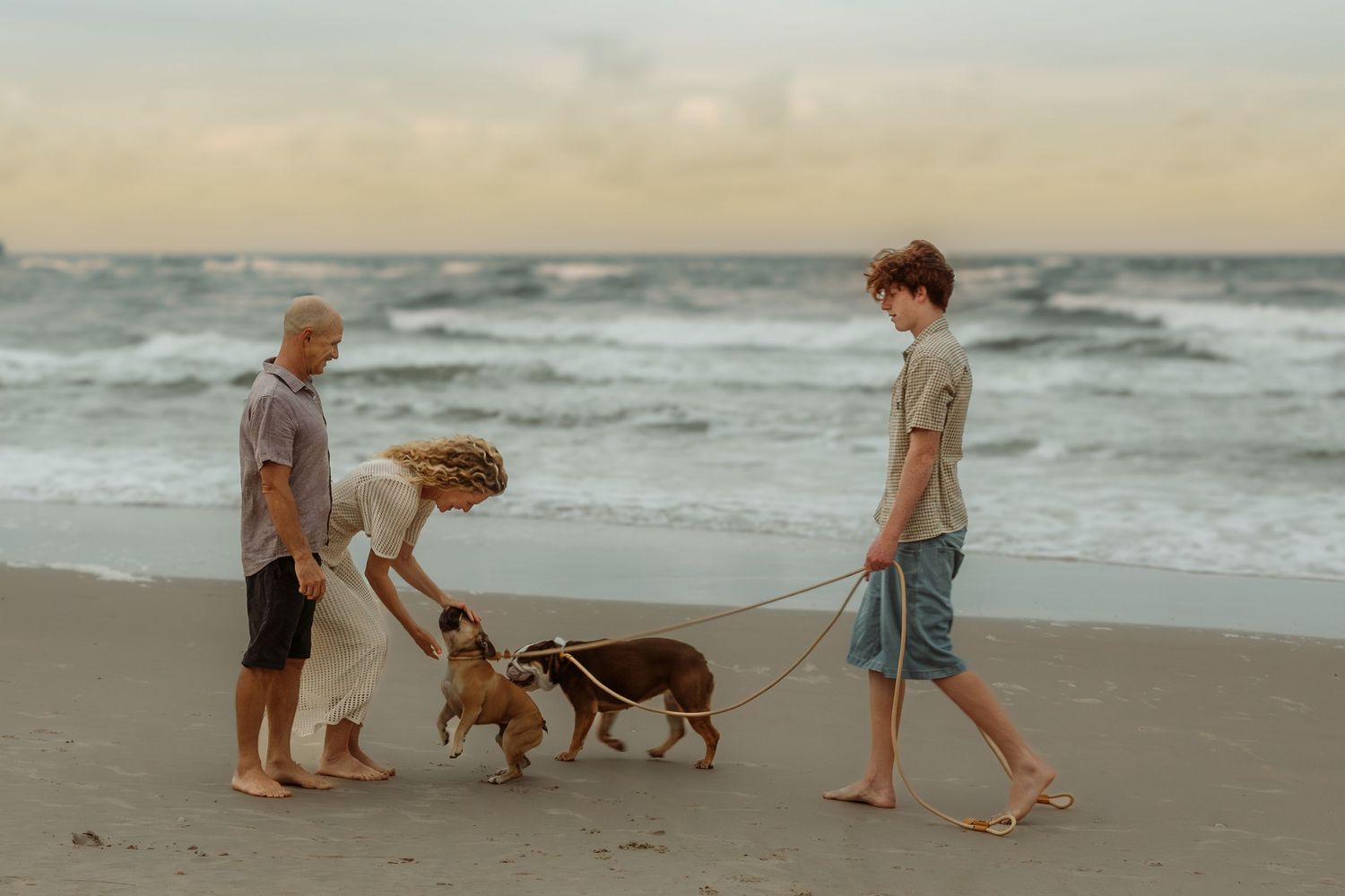 People walking their dog together on a sandy beach at sunset with ocean waves in the background.