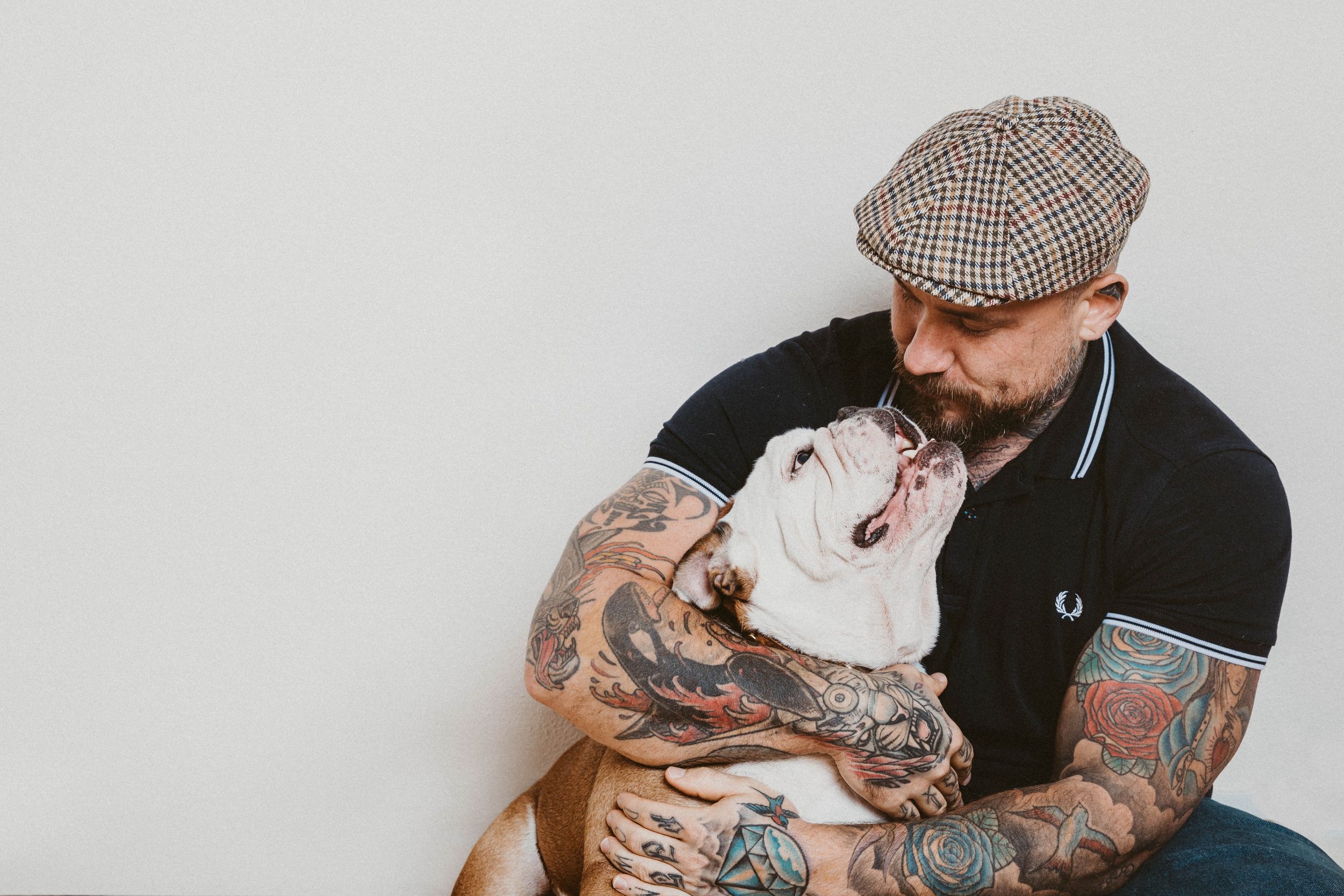 Tattooed person in flat cap holding white pit bull dog showing affection against grey background.