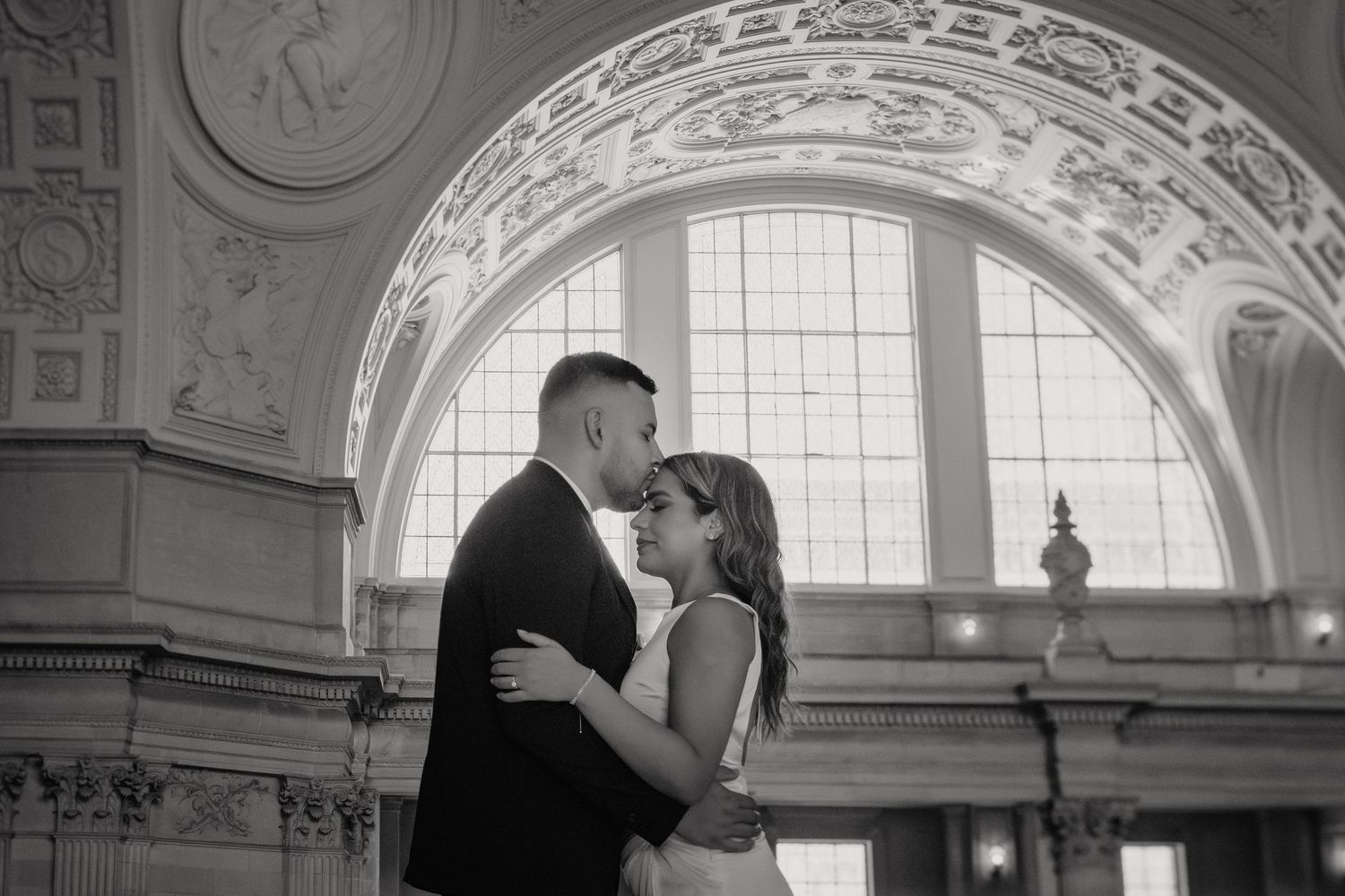 A romantic wedding couple embraces in a grand architectural space with ornate arches and natural window lighting.