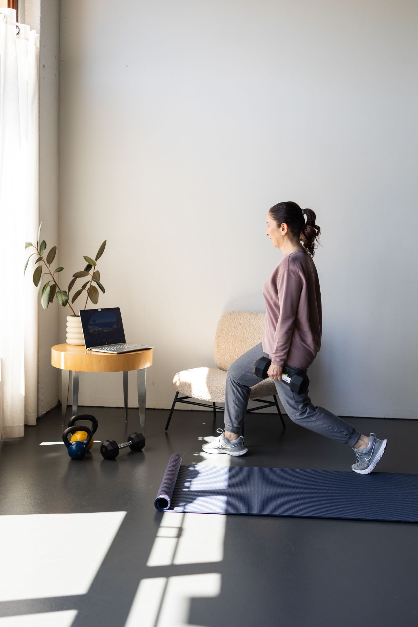 Person doing lunges during home workout session with laptop and exercise equipment in sunlit minimalist room.