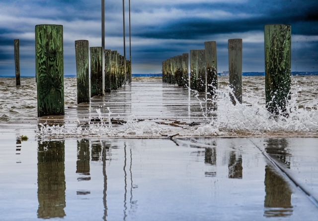 Scenic pier at Colonial Beach, VA with ocean waves crashing and stormy skies, captured behind a waterfront restaurant.