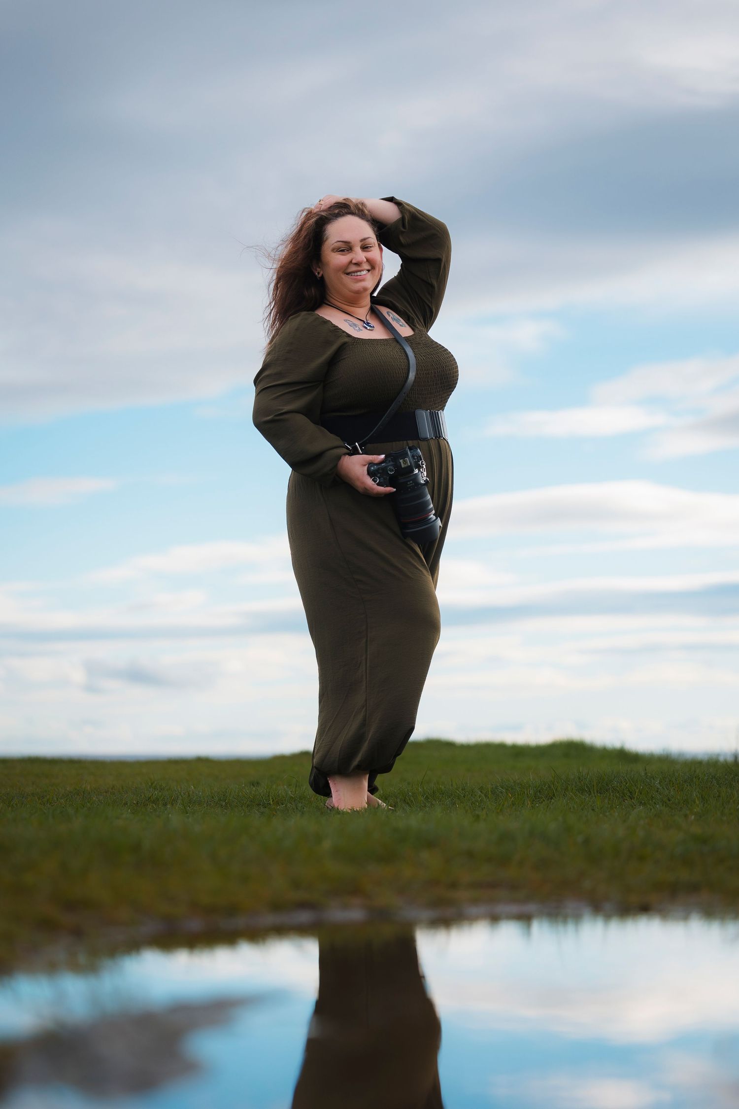 A person in an olive green dress poses confidently by a reflective pool of water in a natural outdoor setting.