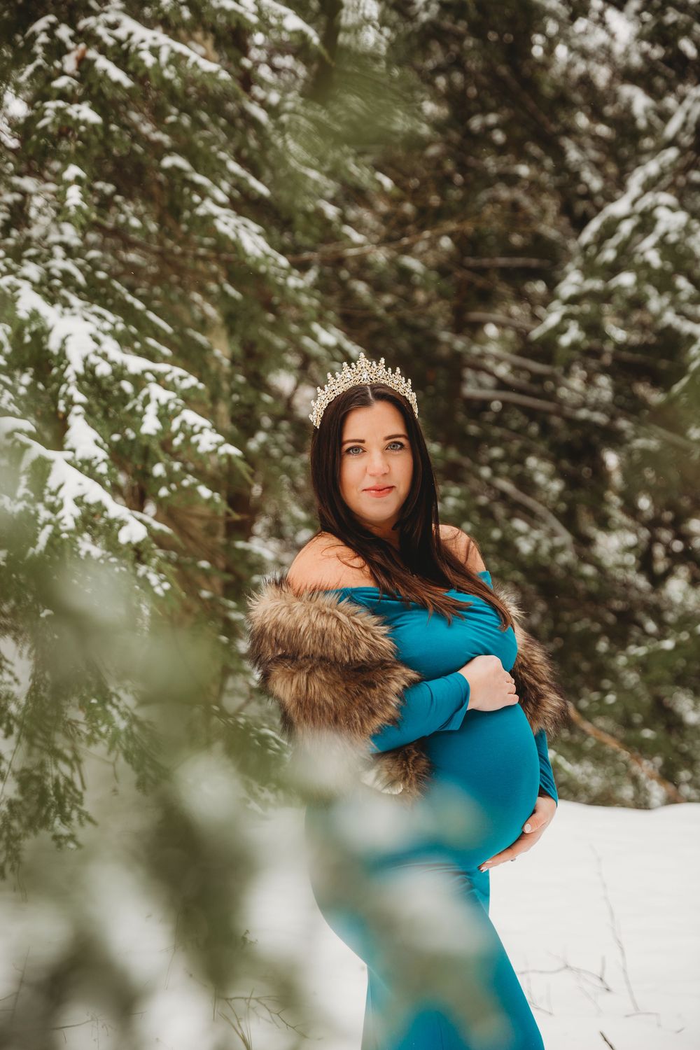 A winter maternity photo featuring a flowing turquoise dress and fur wrap against a snowy evergreen forest backdrop.