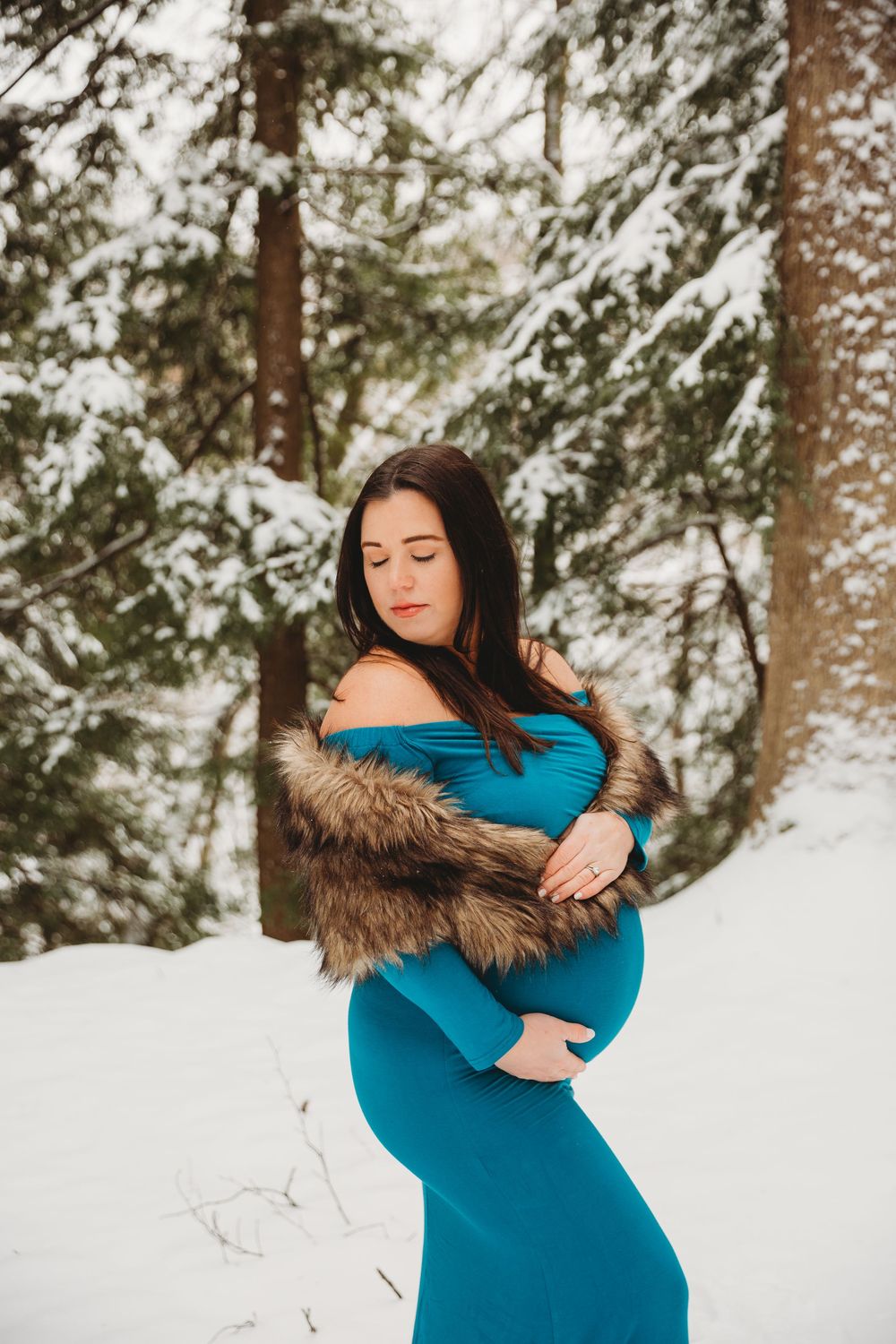 Intimate winter maternity portrait with a turquoise gown amid snow-covered pine trees.