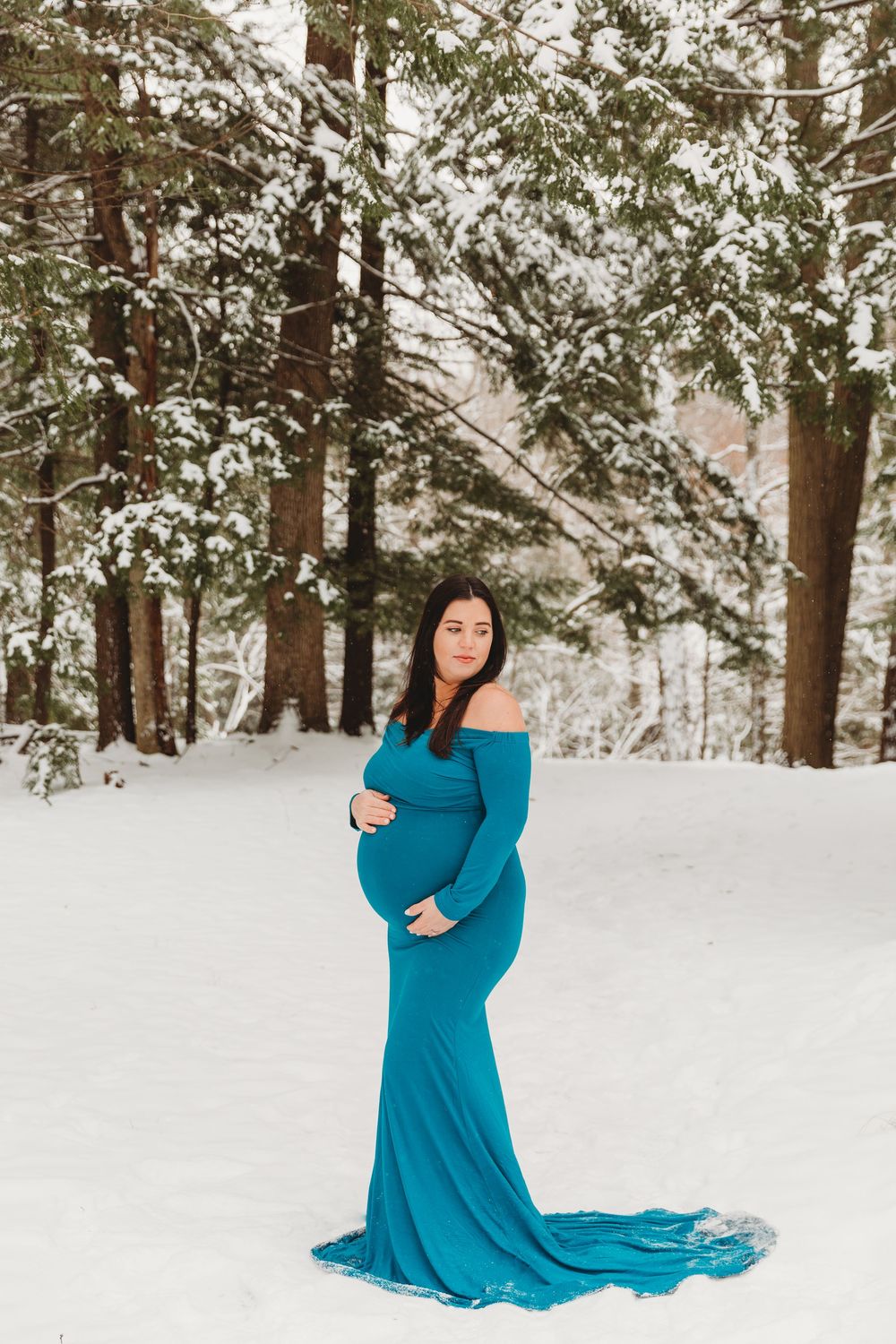 A flowing teal maternity gown creates dramatic movement against a snow-covered forest backdrop.