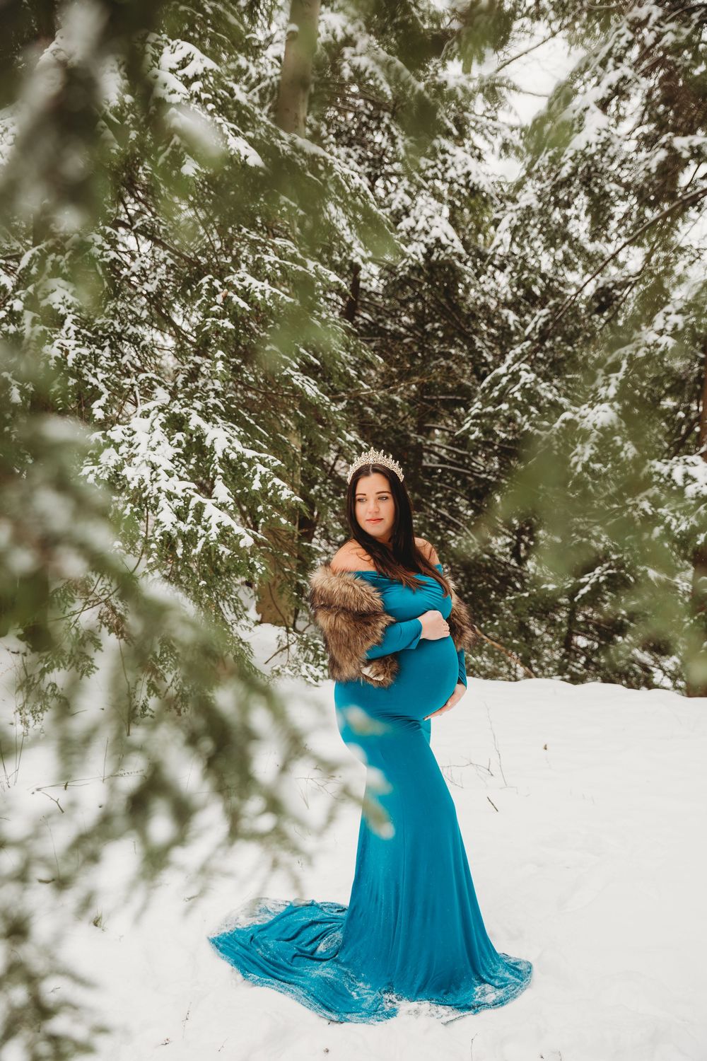 Expectant mother in a vibrant blue gown twirls gracefully in the snow-covered woods.
