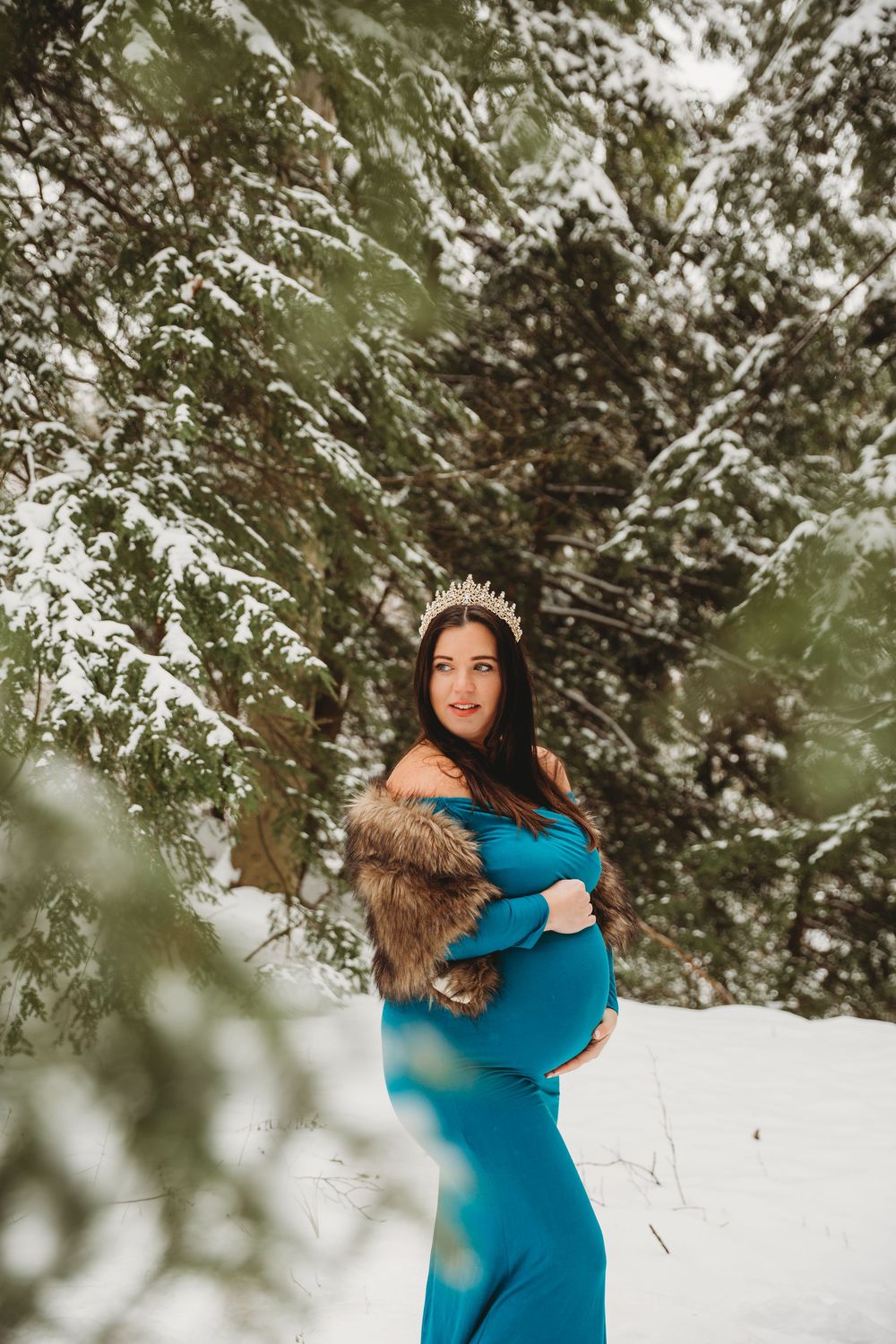 Maternity portrait showing a turquoise dress contrasting beautifully with snow-covered pine trees.