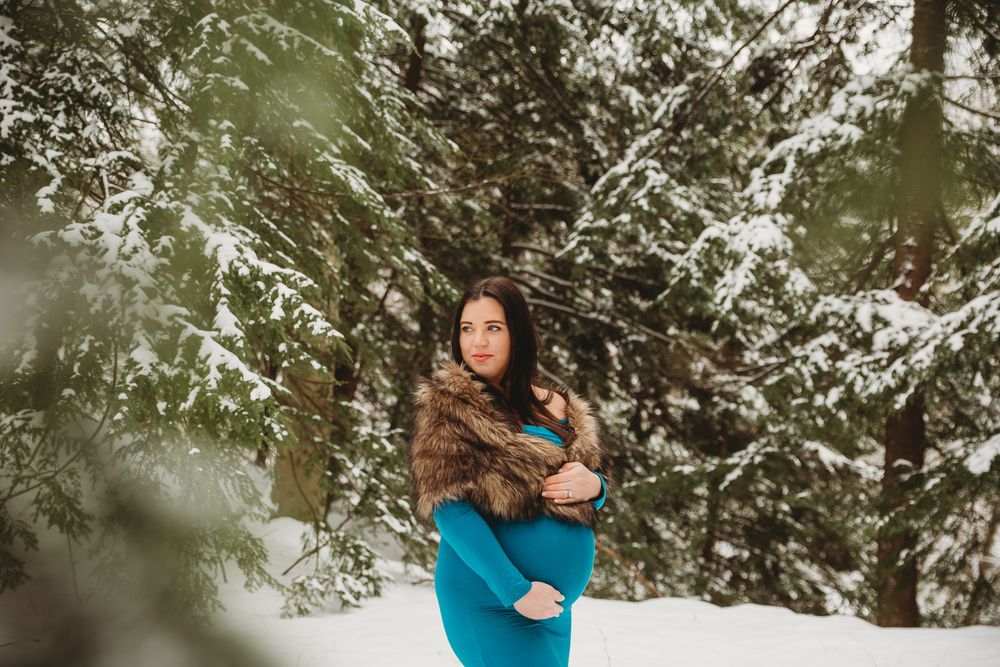 Winter maternity session showcasing an elegant blue dress against snowy forest scenery.