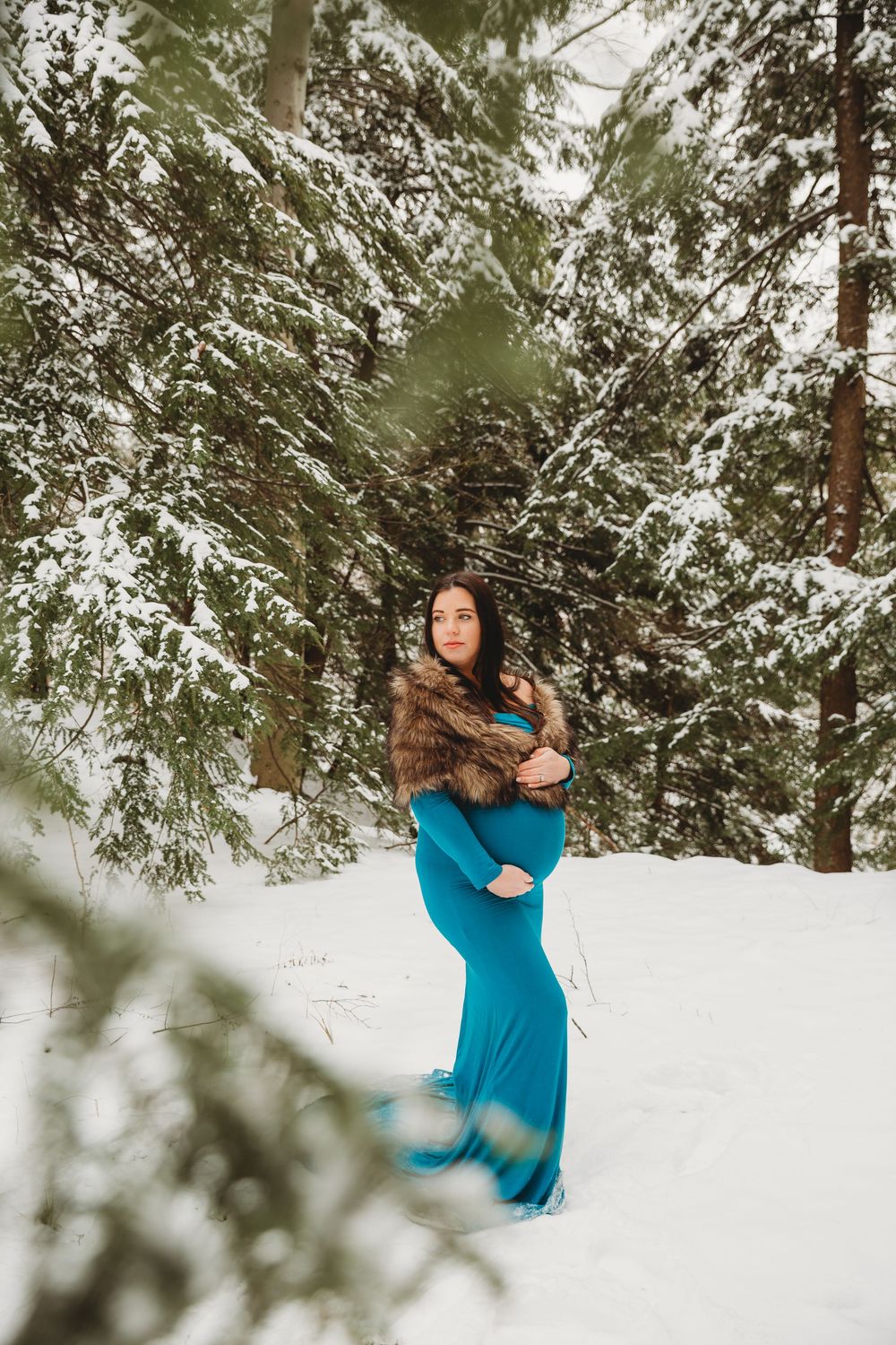 Ethereal winter maternity photo with a flowing teal dress amid snow-dusted evergreen trees.