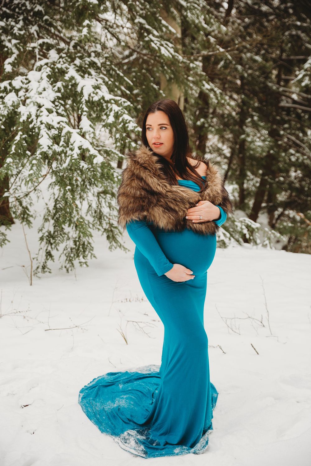 Elegant maternity photo showing a flowing blue dress and fur wrap in a snowy forest setting.