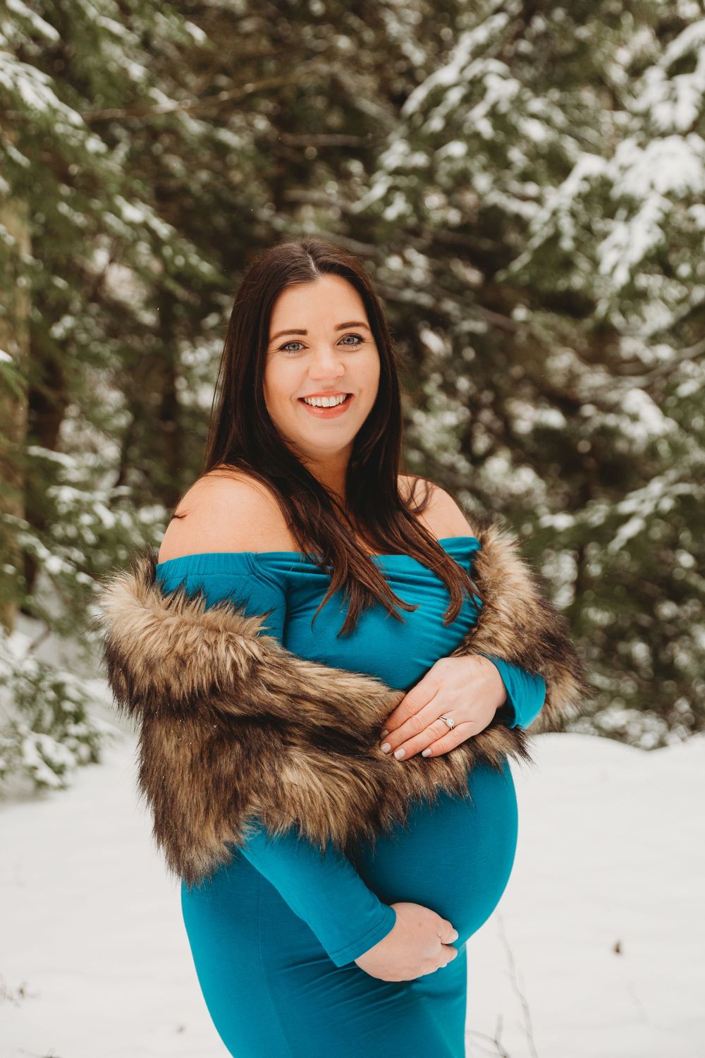 Winter maternity portrait with a fur-trimmed turquoise dress against snowy evergreens.