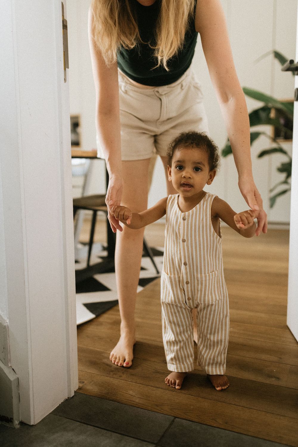 Toddler in striped overalls crawls across a hardwood floor while an adult guides them from behind.