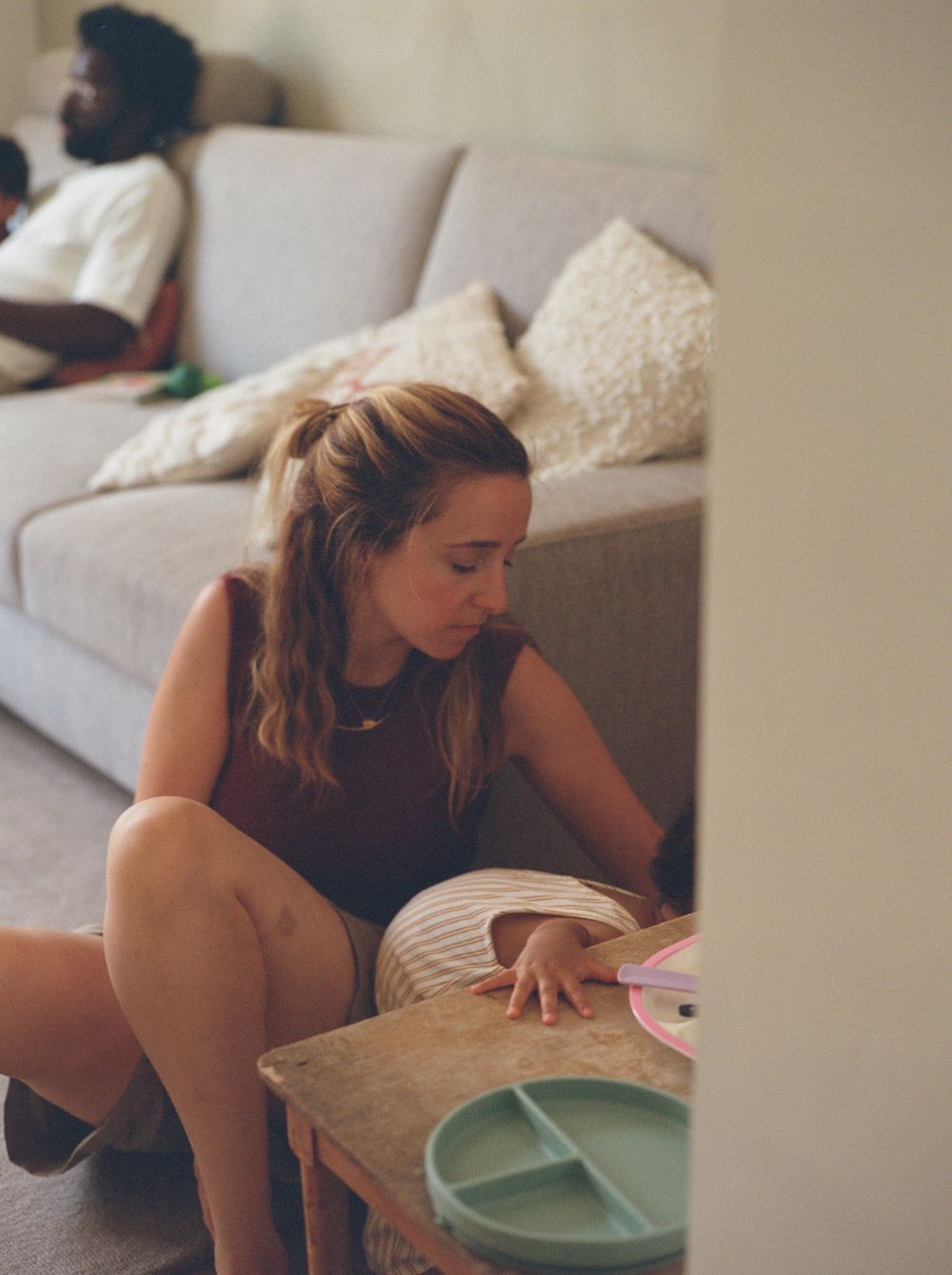 A person in a dark sleeveless top sits on the floor near a gray couch while reaching for items on a coffee table.