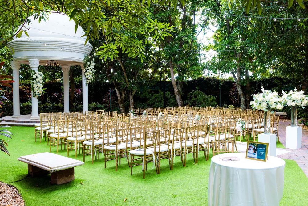 Outdoor garden wedding ceremony setup with white gazebo and wooden chairs on green lawn next to cocktail table.