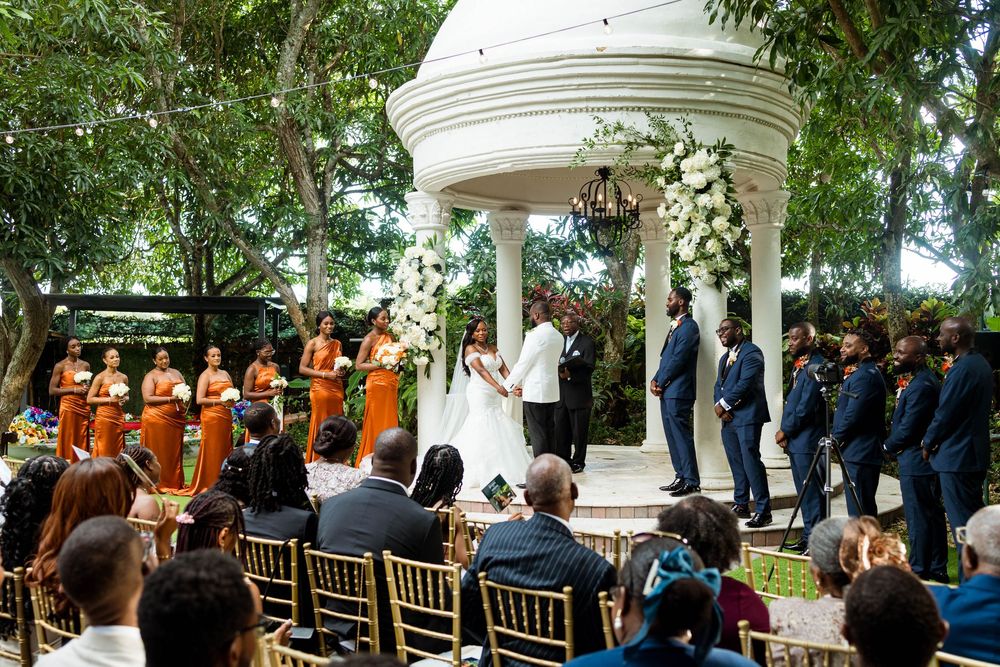 Wedding ceremony at a white gazebo surrounded by bridesmaids in orange dresses and groomsmen in blue suits.