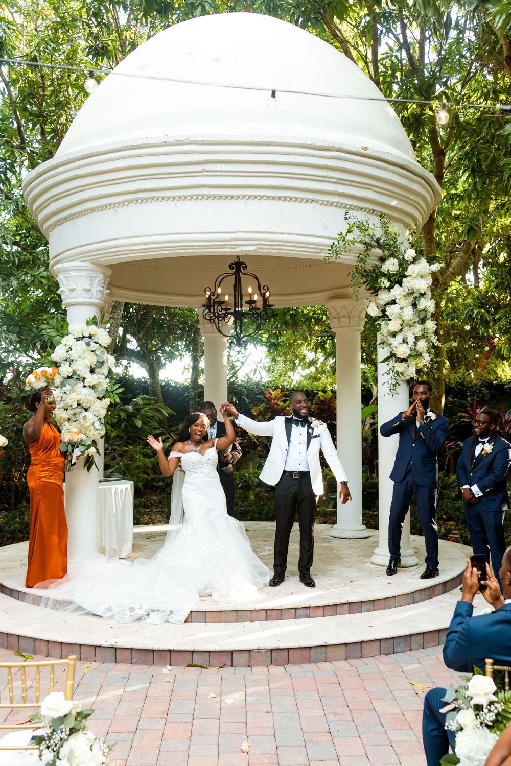 Joyful celebration under a white garden gazebo decorated with elegant floral arrangements for an outdoor wedding ceremony.