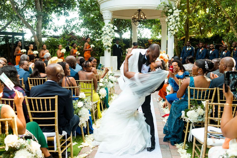 A wedding couple kiss tenderly under a white gazebo as guests celebrate during an outdoor garden ceremony.