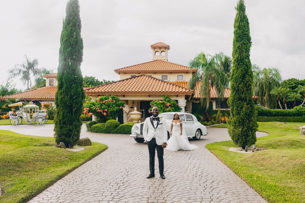 Someone stands on a curved driveway leading to a Mediterranean-style villa with cypress trees and a vintage car in the background.
