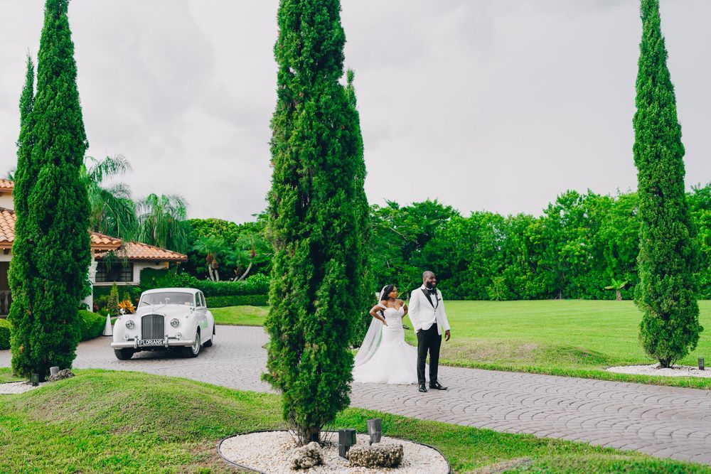 Two people walk along a stone path lined with tall cypress trees near a vintage white car at an elegant venue.