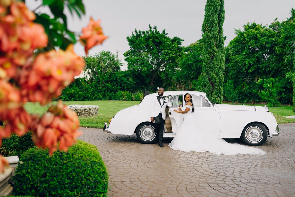 A classic white vintage Rolls-Royce parked on a circular driveway with coral flowers and lush greenery in the background.