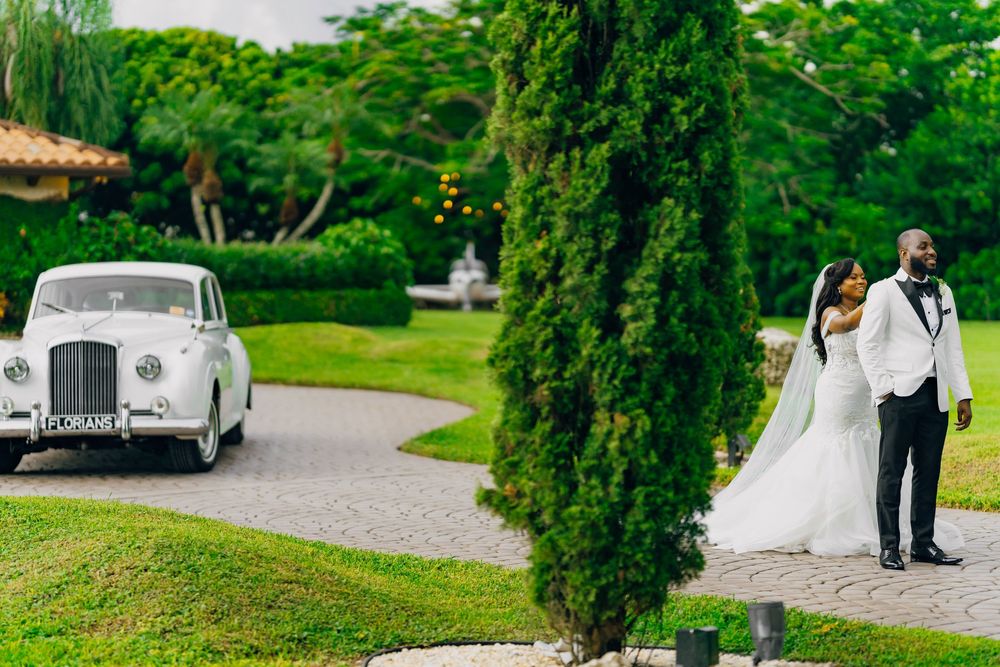 A couple stands together on a garden path while a classic white vintage Bentley car waits in the background.