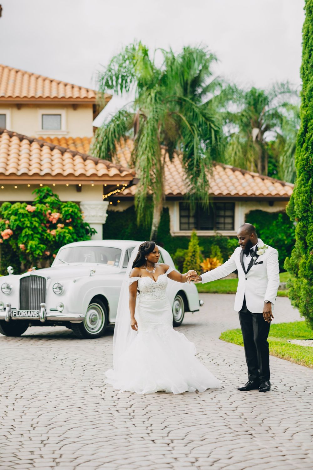 A couple in wedding attire stands by a classic Rolls-Royce on a paved driveway with palm trees and Spanish-style home.
