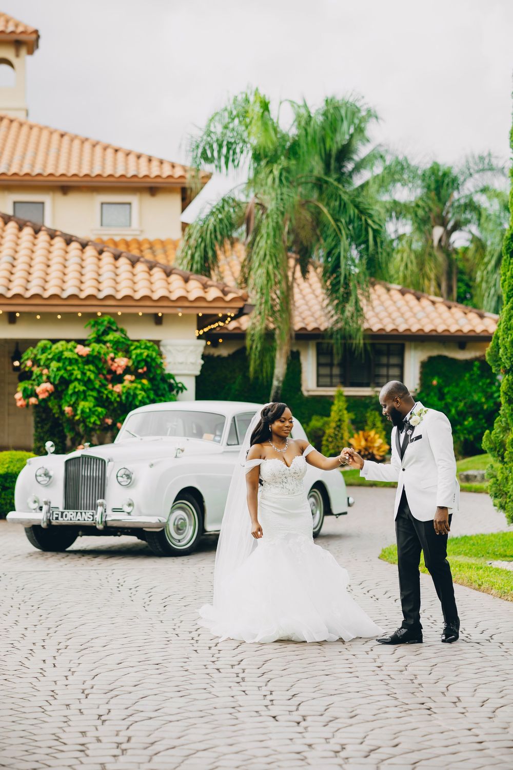 A couple poses in formal wedding attire next to a vintage white car outside a Spanish-style home with palm trees.