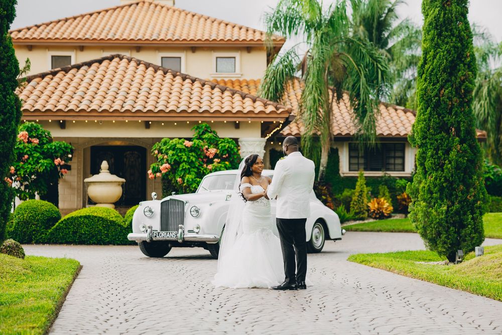 A couple in wedding attire stands beside a vintage white Rolls Royce outside a Spanish-style mansion with palm trees.