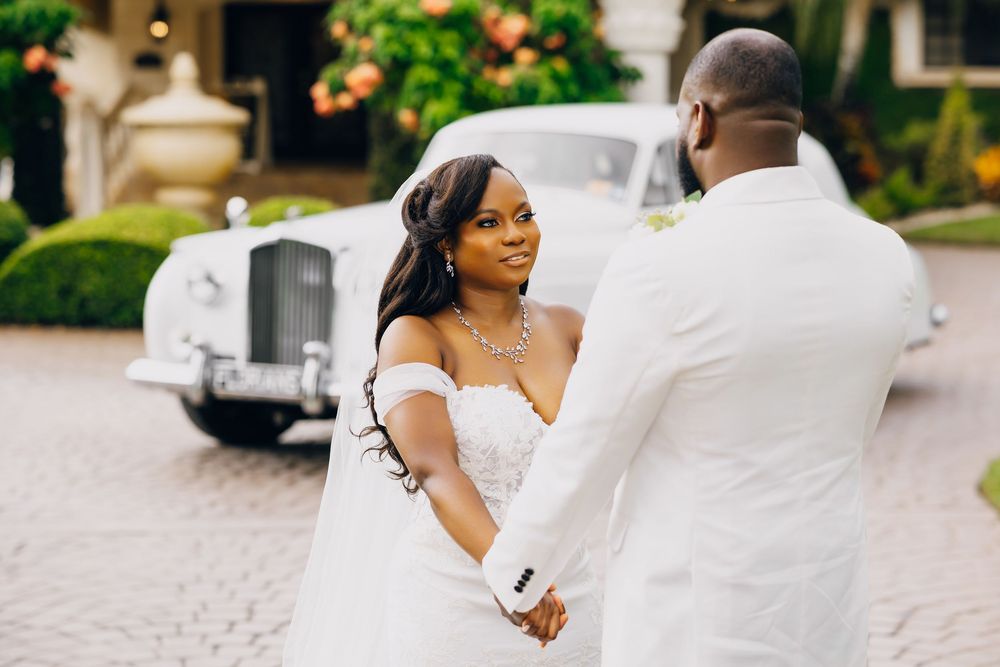 A romantic wedding moment captured outdoors with a white dress and suit against a blurred vintage car background.