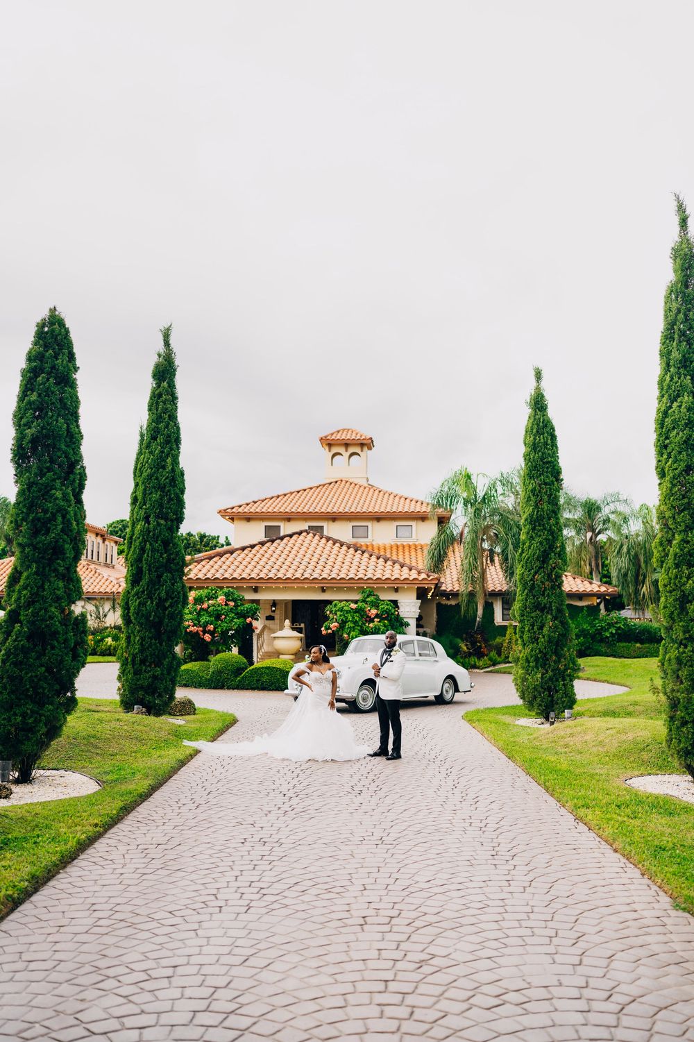 Classic vintage car parked on curved driveway surrounded by tall cypress trees and Mediterranean-style building.