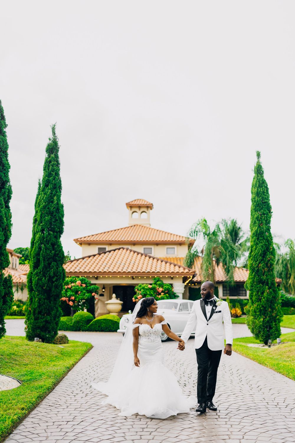 Newlyweds walking down a winding path lined with cypress trees outside a Mediterranean-style building with terracotta roof.