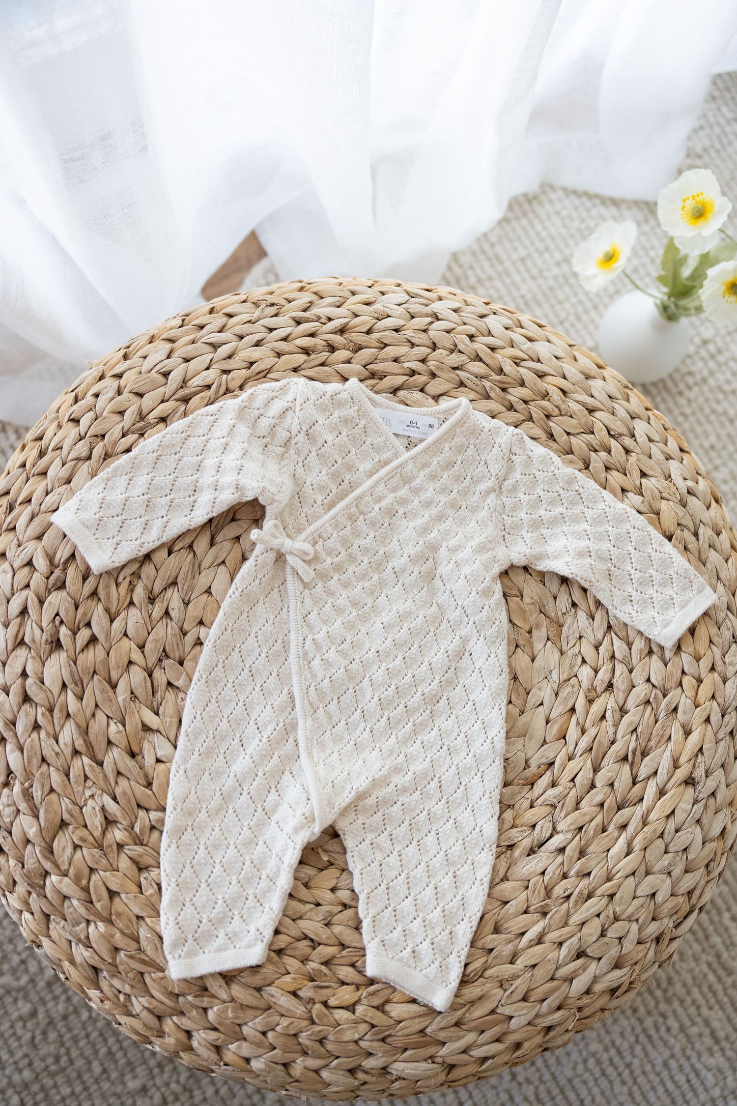 White knitted baby romper displayed on round woven straw mat with small white flowers in background.