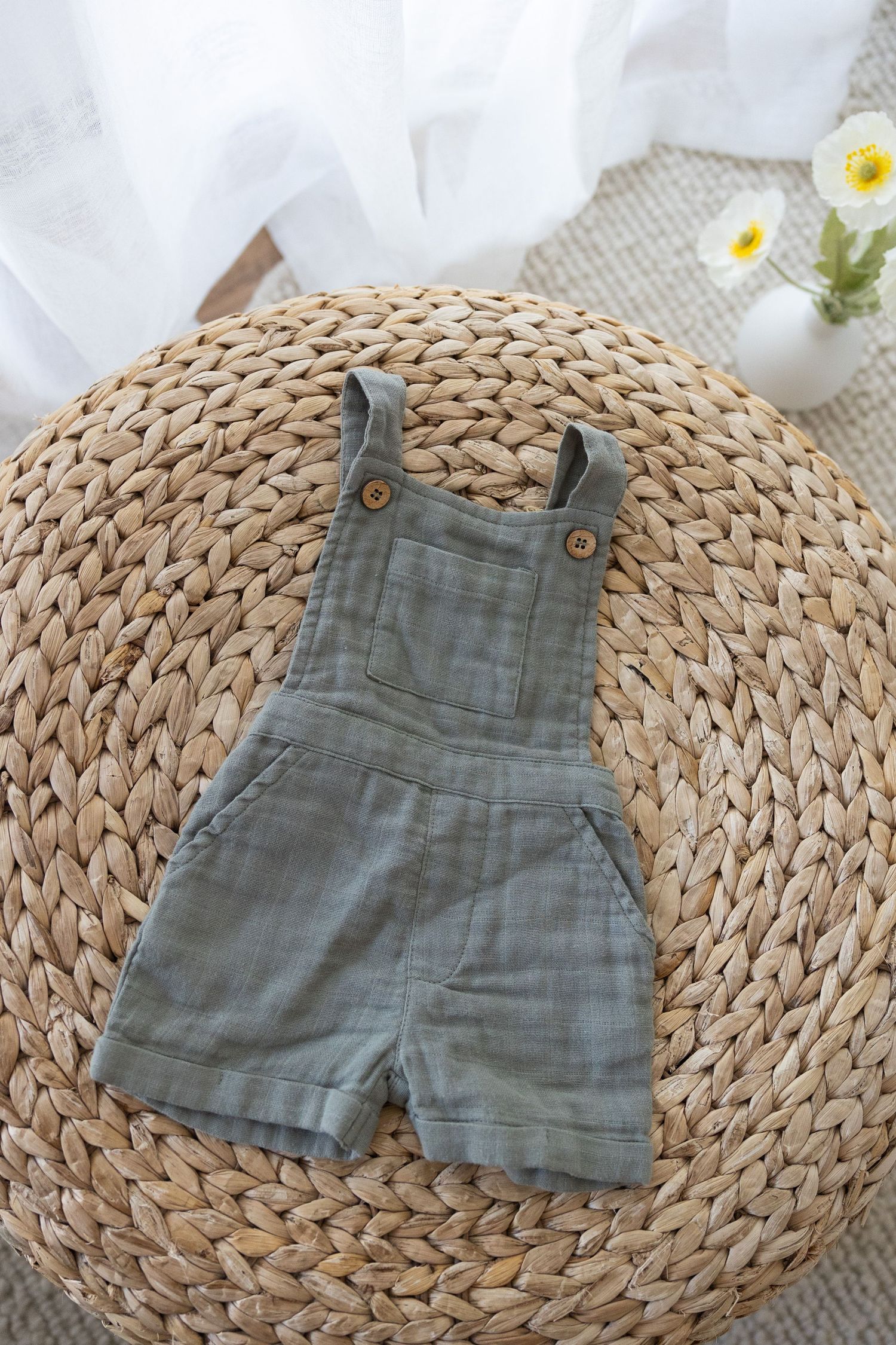 Grey linen baby overalls shorts displayed on round woven straw mat with white fabric and flowers in background.