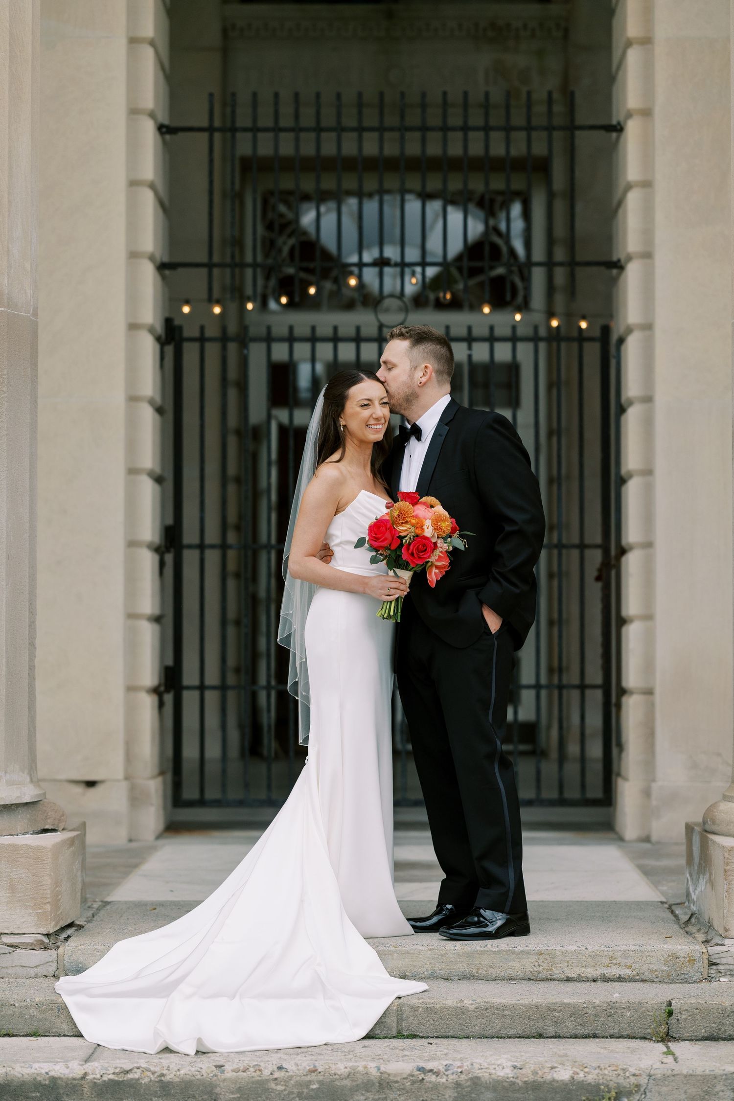 A wedding couple shares a romantic moment on stone steps in front of an ornate iron gate, with coral flowers in bouquet at Hall of Springs.