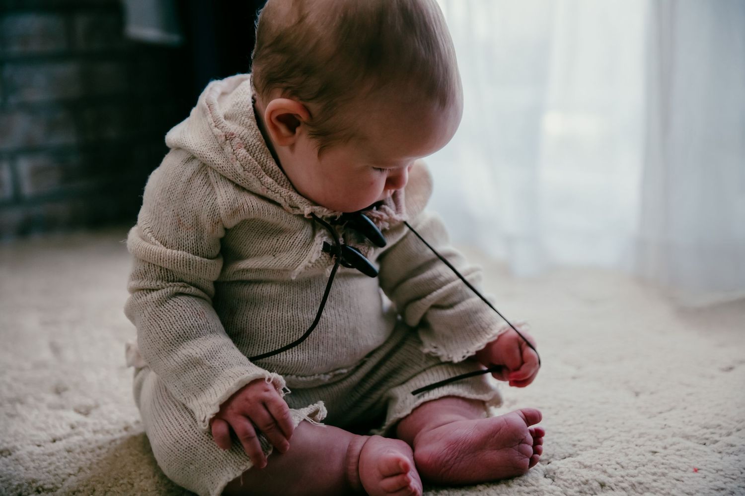 Baby in beige sweater sits on floor playing with piece of string from their top.