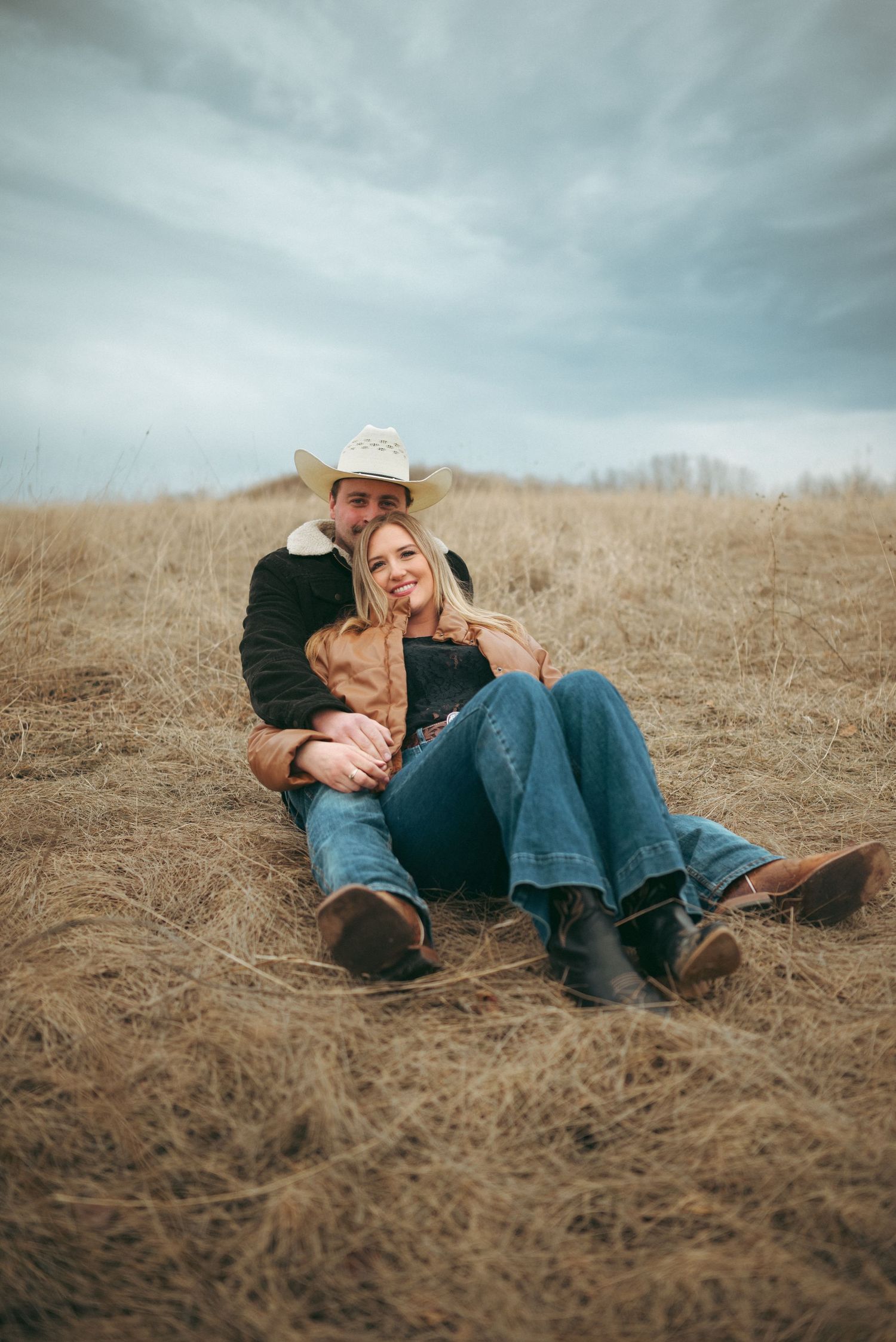 A couple in western attire relaxes together in a field of dry grass under moody skies during an outdoor photoshoot.