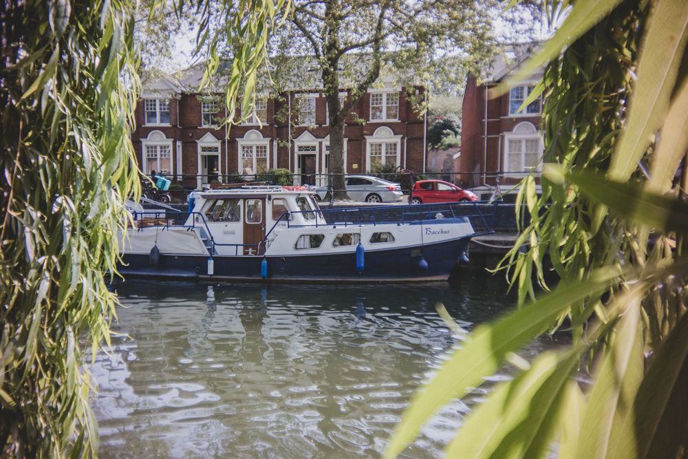 A boat moored in an urban canal with brick townhouses in the background, framed by drooping willow branches.