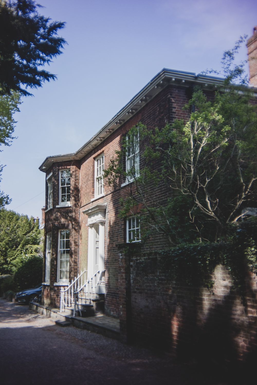Historic brick house with bay windows and white entrance columns surrounded by trees and greenery.