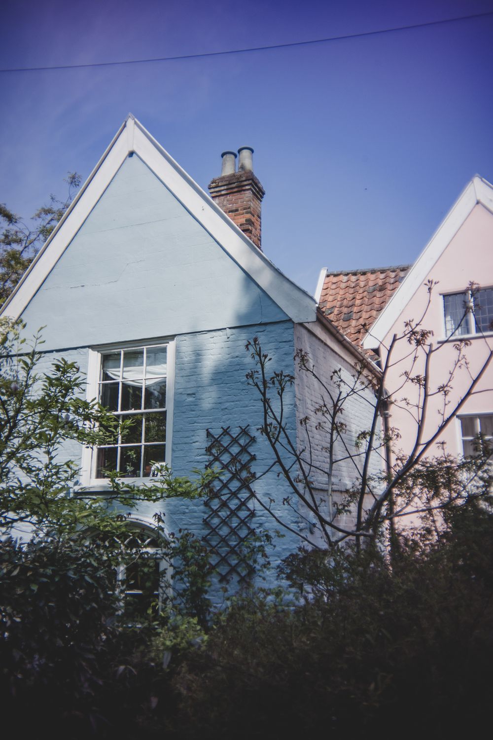 A charming blue cottage with white trim features a gabled roof, brick chimney, and lattice trellis against a pink house next door.