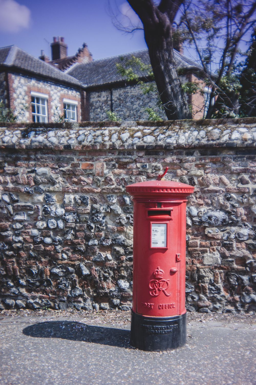 Traditional red British Royal Mail post box mounted against an old stone wall with a house in the background.