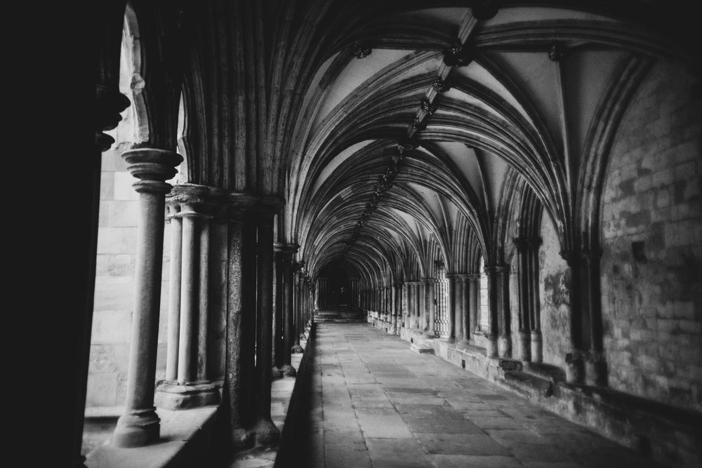 Black and white photograph of a gothic cathedral cloister with vaulted ceilings and stone columns forming a long corridor.