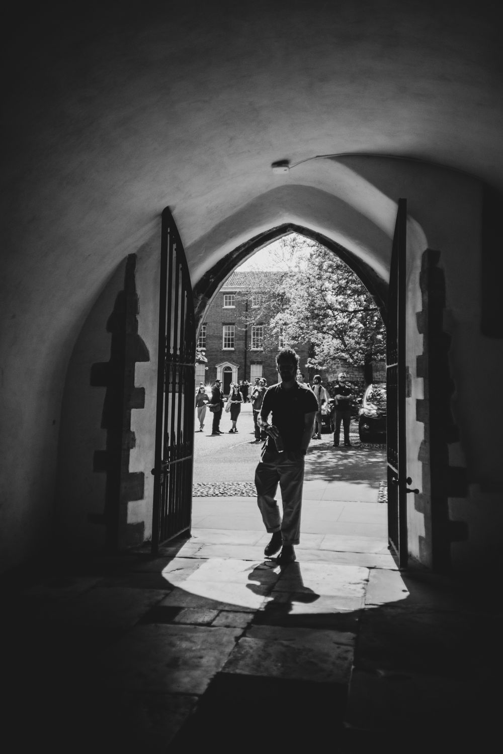 A silhouetted figure walks through a Gothic archway into a sunlit courtyard where people gather in the background.
